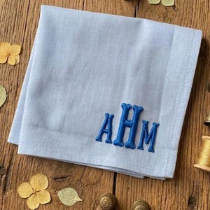 May include: A light blue square handkerchief featuring a centered monogram embroidered in dark blue thread with the letters AHM. The fabric has a visible woven texture and is folded into a neat square shape with clean, straight edges.
