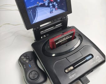 May include: Black Sega Genesis console with a red cartridge labeled "Mega Everdrive" inserted. A small black screen displays a video game scene. A black controller is next to the console. The console is labeled "Mega Drive" and "16 BIT".