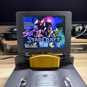 Könnte beinhalten: Eine schwarze Nintendo 64-Konsole mit einer goldenen StarCraft 64-Cartridge. Der Bildschirm zeigt den Titel und die Grafik des Spiels. Die Konsole steht auf einer Holzoberfläche.