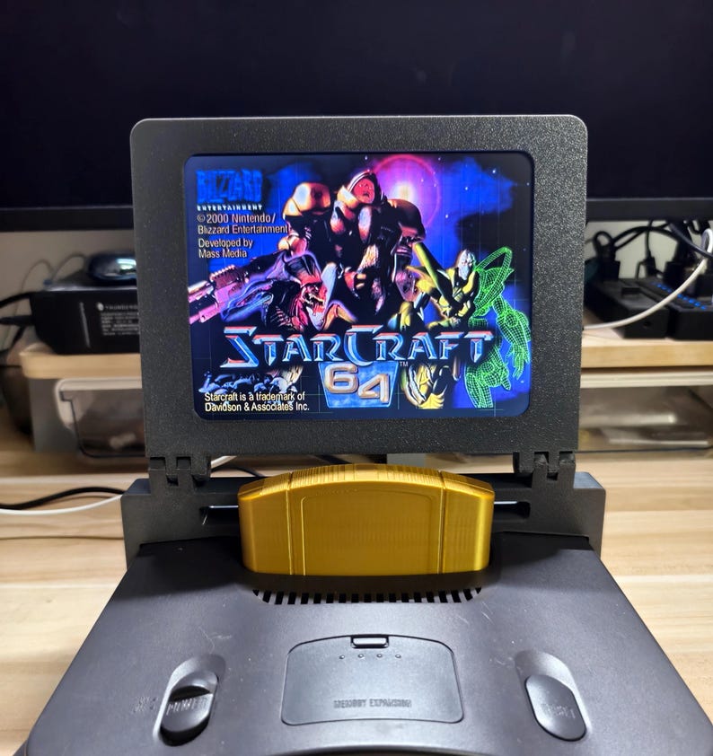 May include: A black Nintendo 64 console with a gold StarCraft 64 cartridge inserted. The screen displays the game's title and graphics. The console is on a wooden surface.