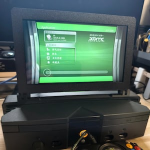 Może przedstawiać: Czarna konsola Xbox z ekranem wyświetlającym zielony interfejs. Ekran jest zamontowany na g&oacute;rze konsoli. Na ekranie wyświetla się słowo "Applications" i inny tekst w obcym języku. Czarny kontroler znajduje się na pierwszym planie.
