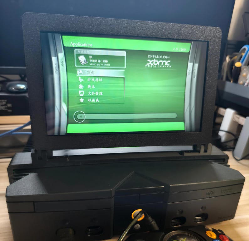 Może przedstawiać: Czarna konsola Xbox z ekranem wyświetlającym zielony interfejs. Ekran jest zamontowany na g&oacute;rze konsoli. Na ekranie wyświetla się słowo "Applications" i inny tekst w obcym języku. Czarny kontroler znajduje się na pierwszym planie.