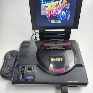 May include: Black Sega Genesis Mega Drive console with a screen displaying the "Final Fight" game. A red Everdrive cartridge is inserted. A black controller is on the left. The console has a "16-BIT" label and "Mega Drive" text.