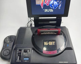 May include: Black Sega Genesis Mega Drive console with a screen displaying the "Final Fight" game. A red Everdrive cartridge is inserted. A black controller is on the left. The console has a "16-BIT" label and "Mega Drive" text.