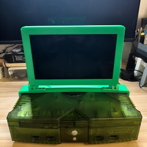 Könnte beinhalten: Eine transparente grüne Spielkonsole mit einem passenden grünen Bildschirm. Die Konsole hat eine rechteckige Form mit mehreren Tasten und Anschlüssen. Der Bildschirm ist aufrecht und an der Konsole befestigt.