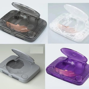 May include: Four transparent plastic cases, each resembling a classic video game console with an open lid. The cases are in shades of grey, white, and purple. The cases have a circular button on the front.