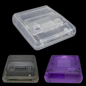 May include: Clear plastic protective cases for a Super Nintendo console. The image shows three cases: one clear, one gray, and one purple. The cases are rectangular with slots for game cartridges and controller ports. The cases are designed to protect the console from dust and damage.