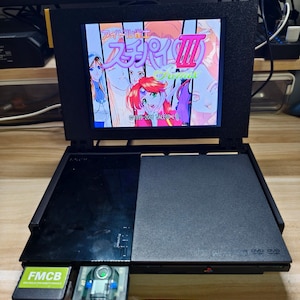 Puede incluir: Consola PlayStation 2 negra con una pantalla que muestra texto japonés y personajes de estilo anime. La consola tiene una unidad de DVD/DVD-ROM y una ranura para tarjetas de memoria. Una tarjeta "FMCB" verde y blanca está conectada.