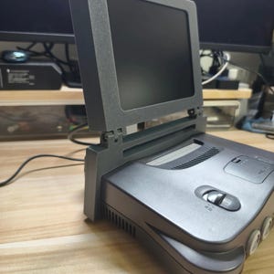 May include: A gray Nintendo 64 console with a matching screen attachment. The screen is black, and the console has a power button and cartridge slot. The setup is on a light-colored surface.