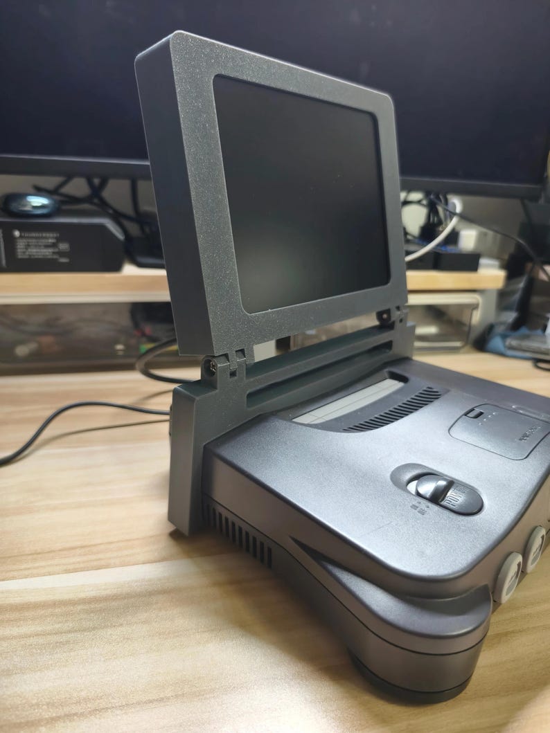 May include: A gray Nintendo 64 console with a matching screen attachment. The screen is black, and the console has a power button and cartridge slot. The setup is on a light-colored surface.