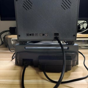 May include: A gray retro gaming console with a screen, vents, and various ports. The console is connected to a power supply and other devices via black cables. The setup is on a light-colored wooden surface.