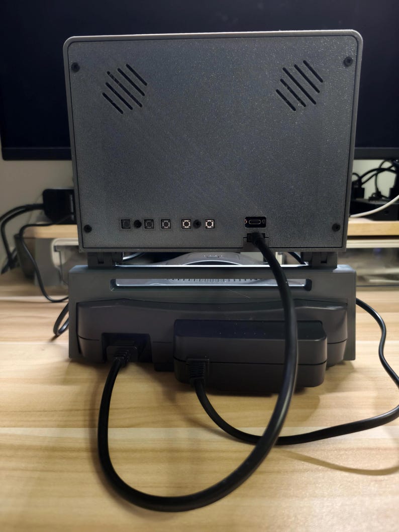 May include: A gray retro gaming console with a screen, vents, and various ports. The console is connected to a power supply and other devices via black cables. The setup is on a light-colored wooden surface.