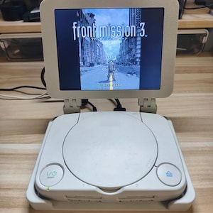 May include: A white PlayStation console with a screen displaying the game "front mission 3." The screen is attached to the console via hinges. The console has a power button, a reset button, and two controller ports.