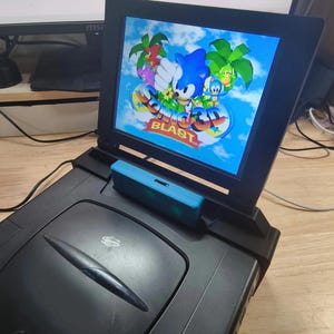 May include: A black portable video game console with a screen showing a Sonic the Hedgehog game. The screen displays Sonic, Tails, and other characters on a tropical island. A blue game cartridge is inserted.