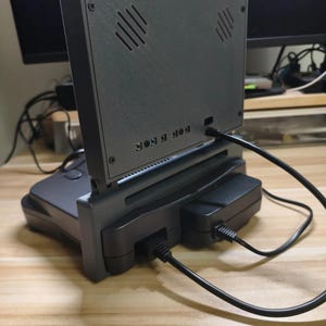 May include: A gray retro gaming console with a screen attached. The screen has diagonal vents and is connected to the console with a stand. Black cables are plugged into the console and a black power adapter. The console is on a wooden surface.