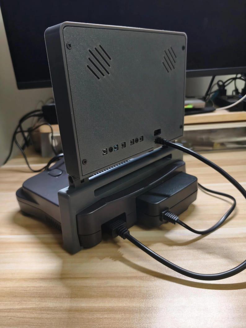May include: A gray retro gaming console with a screen attached. The screen has diagonal vents and is connected to the console with a stand. Black cables are plugged into the console and a black power adapter. The console is on a wooden surface.