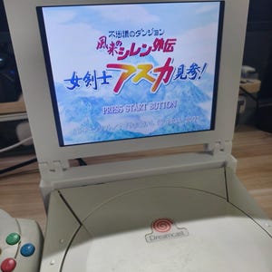 May include: A white Dreamcast console with a screen displaying Japanese text. The screen reads "PRESS START BUTTON" and features colorful Japanese characters. The console has a "Dreamcast" logo.