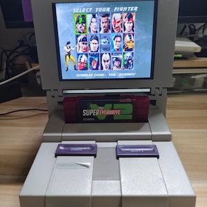 May include: A grey Super Nintendo console with a screen showing the "Select Your Fighter" screen from a fighting game. The game cartridge reads "Super Everdrive V2". The console has purple power and reset buttons.