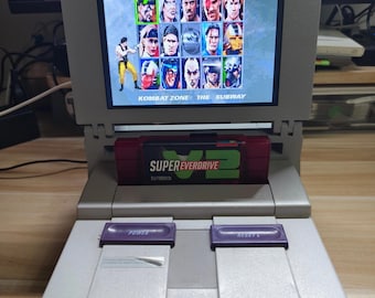 Puede incluir: Una consola Super Nintendo gris con una pantalla que muestra la pantalla "Select Your Fighter" de un juego de lucha. El cartucho del juego dice "Super Everdrive V2". La consola tiene botones de encendido y reinicio morados.