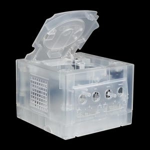 May include: A clear, transparent plastic video game console with the lid open. The console is a cube shape with a grid vent on the side. The front panel has circular ports and buttons. The lid is also transparent, showing the internal components.