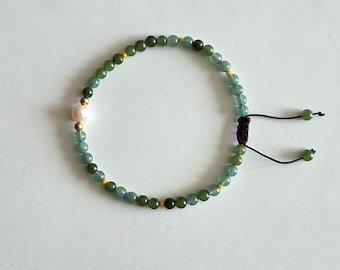 May include: A beaded bracelet featuring green and dark green beads, a single pink pearl, and gold-colored accents. The bracelet has an adjustable closure with black cords and green beads at the ends.