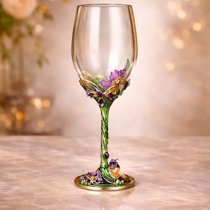 Puede incluir: Una copa de vino transparente con base y tallo decorativos. La base y el tallo están pintados de verde con detalles florales morados y ribete dorado. La copa está vacía y se apoya sobre una superficie de mármol.