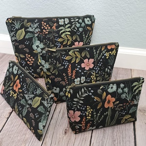 May include: Four black zippered pouches of varying sizes, each adorned with a floral pattern. The design features a dark background with colorful flowers and foliage in shades of pink, orange, and blue. The pouches have a rectangular shape with a slight trapezoid design.
