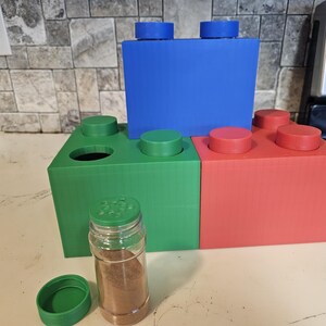 May include: A set of three spice jars made to look like building blocks. The jars are green, blue, and red. The jars are arranged in a stack with the green jar on the bottom, the blue jar in the middle, and the red jar on top. A small green lid and a glass spice jar with a green lid are in the foreground.