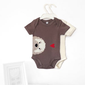 May include: Two baby onesies, one brown with a white dog face and red heart applique, and one white. Both have short sleeves and snap closures.