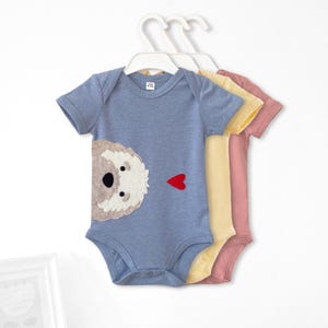 May include: Three baby onesies with short sleeves. The onesies are blue, yellow, and pink. Each onesie has a felt applique of a dog's face with a red heart on the front.