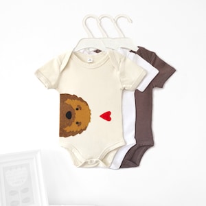 May include: Three baby onesies, one with a brown felt dog applique and a red felt heart, one white, and one brown.