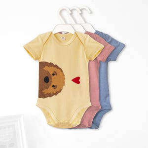 May include: Three baby onesies, one yellow with a brown dog face and a red heart, one pink, and one blue.