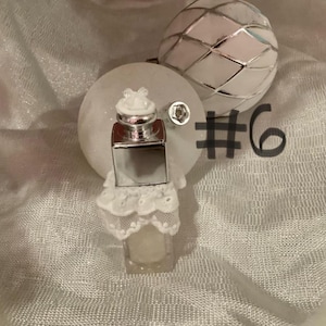 May include: A small, clear perfume bottle with a silver cap and a white lace base. The bottle is next to a white, round ornament and a decorative, silver-colored sphere. The number 6 is written in black.