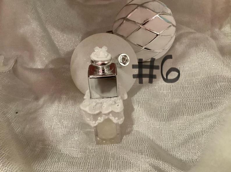May include: A small, clear perfume bottle with a silver cap and a white lace base. The bottle is next to a white, round ornament and a decorative, silver-colored sphere. The number 6 is written in black.