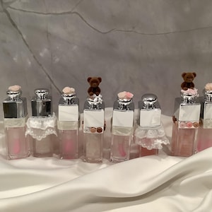 May include: A collection of lip gloss bottles with silver caps and pink liquid. Some bottles are adorned with small teddy bear figurines and pink flowers. The bottles are arranged on a white satin surface.