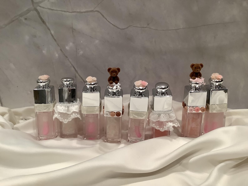 May include: A collection of lip gloss bottles with silver caps and pink liquid. Some bottles are adorned with small teddy bear figurines and pink flowers. The bottles are arranged on a white satin surface.