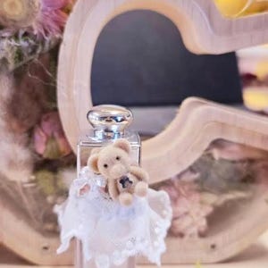 May include: A clear rectangular lip gloss bottle with a silver cap, adorned with a small teddy bear wearing a white lace dress. The background includes a wooden heart-shaped decoration and soft lighting. Text at the top reads "Lip Gloss March 8".