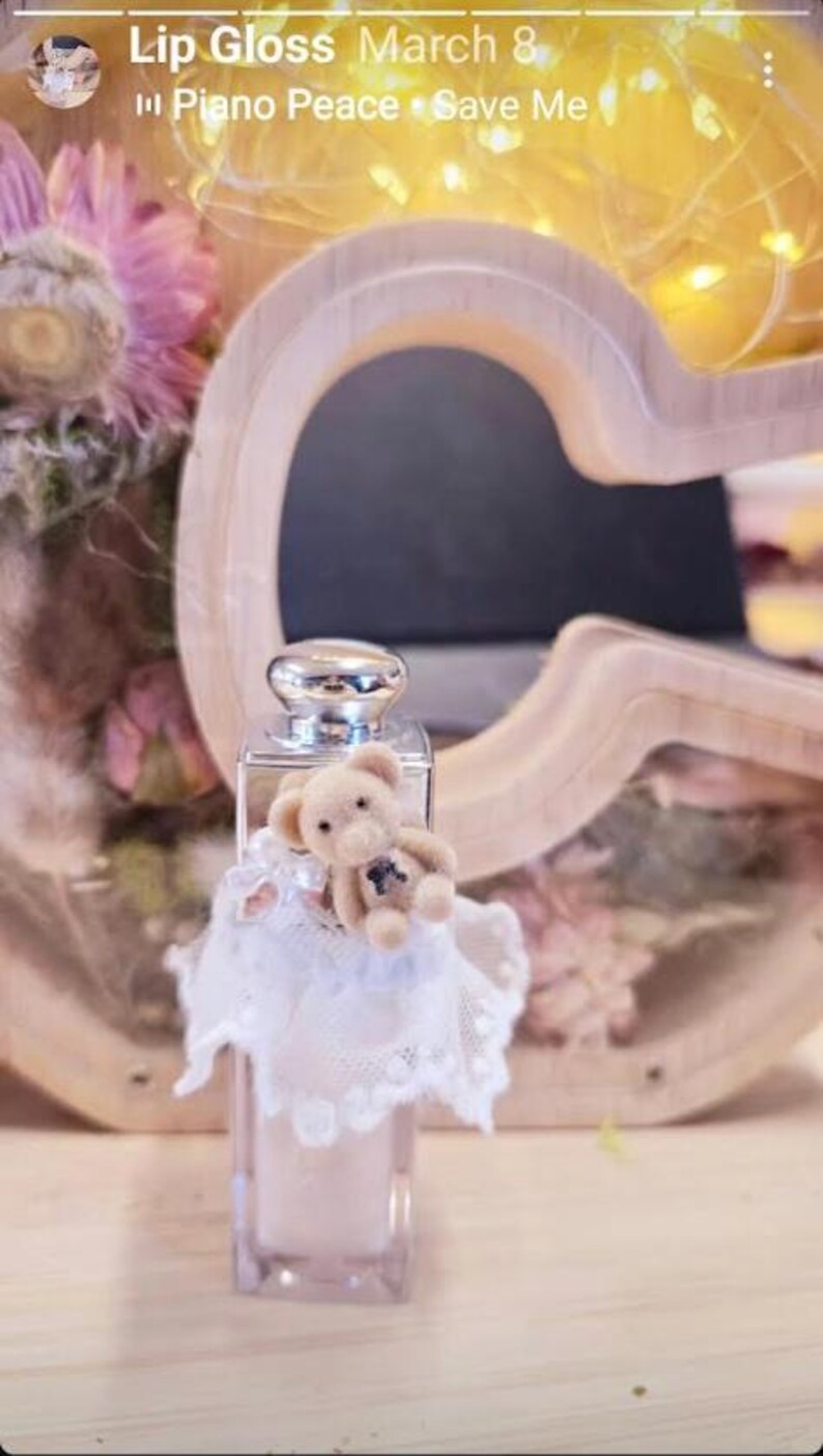 May include: A clear rectangular lip gloss bottle with a silver cap, adorned with a small teddy bear wearing a white lace dress. The background includes a wooden heart-shaped decoration and soft lighting. Text at the top reads "Lip Gloss March 8".