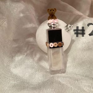 May include: A decorative ornament featuring a small teddy bear atop a perfume bottle-shaped design. The bottle has a black top and is adorned with gold and bronze-colored sequins. The ornament is set against a white, textured background.