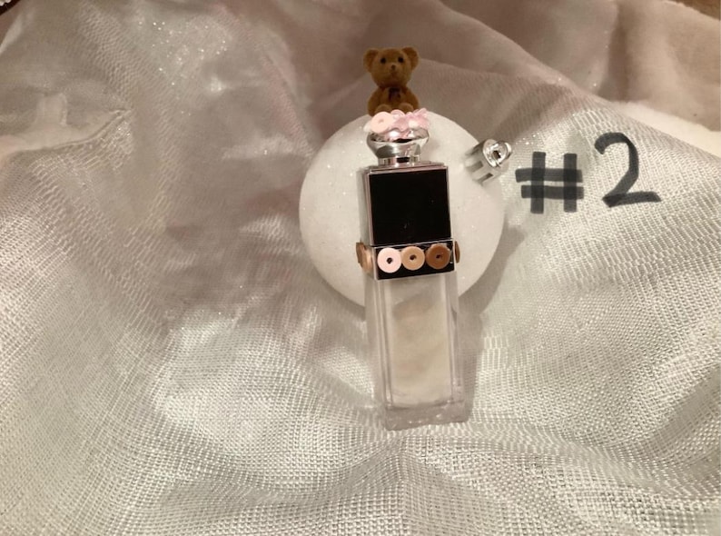May include: A decorative ornament featuring a small teddy bear atop a perfume bottle-shaped design. The bottle has a black top and is adorned with gold and bronze-colored sequins. The ornament is set against a white, textured background.