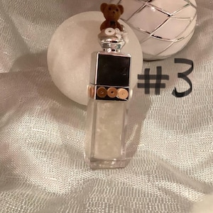 May include: A clear, rectangular perfume bottle with a silver cap, adorned with a small brown teddy bear. The bottle has a black square on the front and gold accents. A white sphere and a decorative silver and white ball are in the background. The number 3 is written in black.