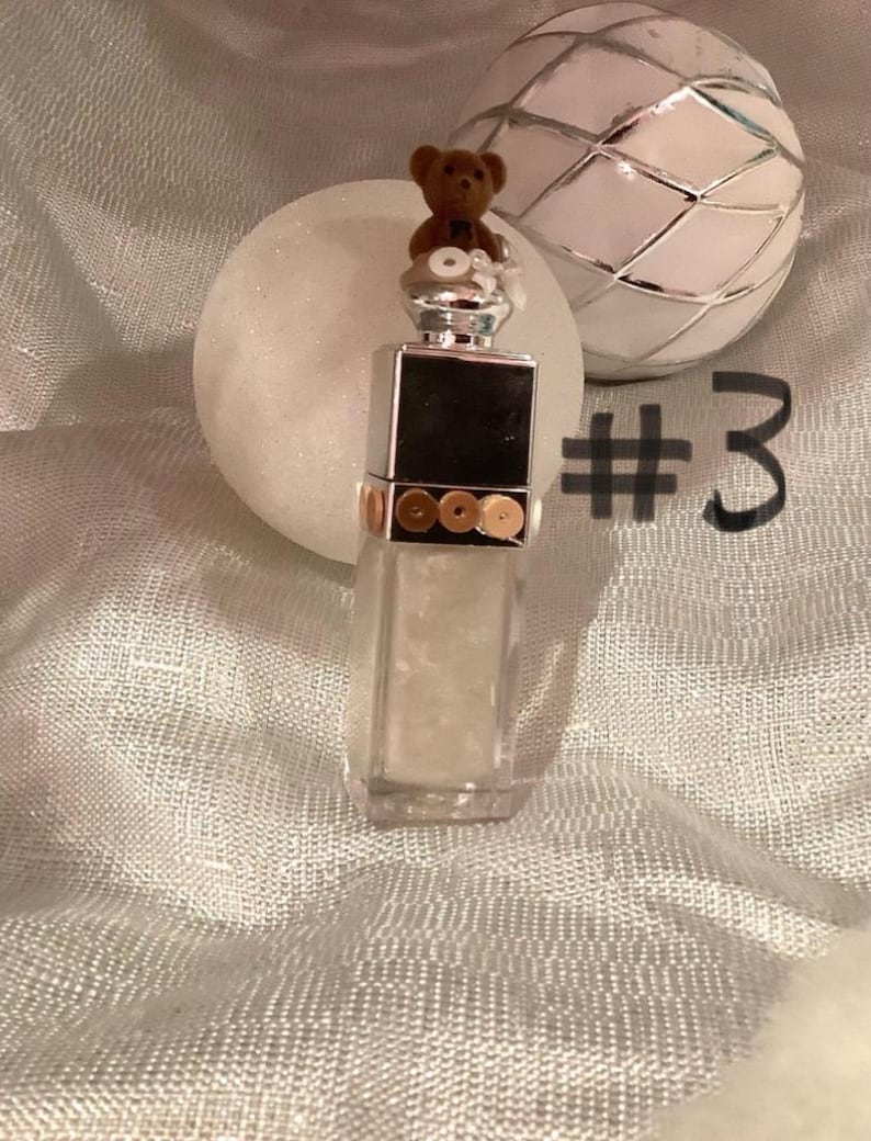 May include: A clear, rectangular perfume bottle with a silver cap, adorned with a small brown teddy bear. The bottle has a black square on the front and gold accents. A white sphere and a decorative silver and white ball are in the background. The number 3 is written in black.