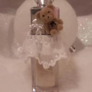 May include: A clear, rectangular lip gloss bottle with a silver cap, adorned with a small teddy bear and a white lace skirt. The bottle is filled with a light-colored gloss. The text "Lip Gloss" and "March 8" are visible at the top.