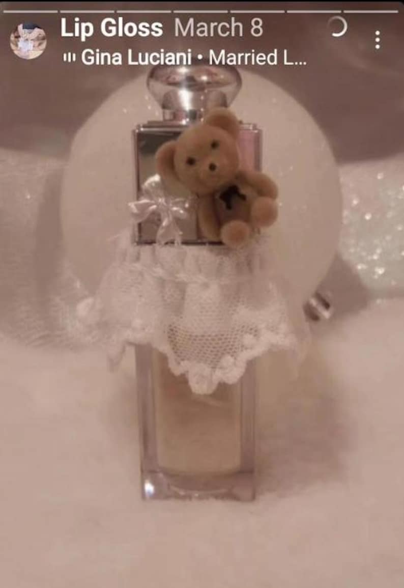 May include: A clear, rectangular lip gloss bottle with a silver cap, adorned with a small teddy bear and a white lace skirt. The bottle is filled with a light-colored gloss. The text "Lip Gloss" and "March 8" are visible at the top.