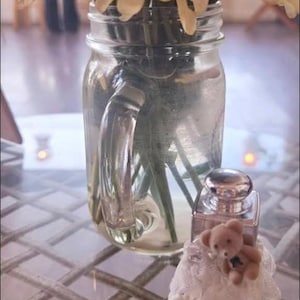 May include: A clear glass jar with a handle holds a bouquet of cream-colored flowers. A small perfume bottle with a silver cap and a teddy bear figurine sits in front of the jar. The items are on a table with a patterned surface.