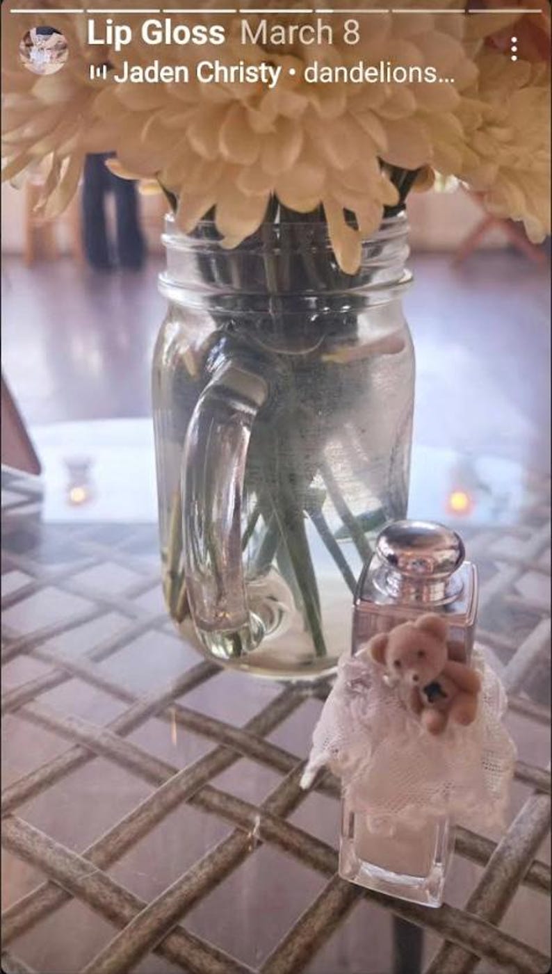 May include: A clear glass jar with a handle holds a bouquet of cream-colored flowers. A small perfume bottle with a silver cap and a teddy bear figurine sits in front of the jar. The items are on a table with a patterned surface.