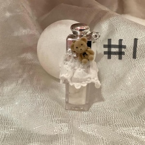 May include: A small, clear perfume bottle with a silver cap and a tiny teddy bear attached. The bottle is adorned with a white lace collar. The background is a soft, textured white fabric.