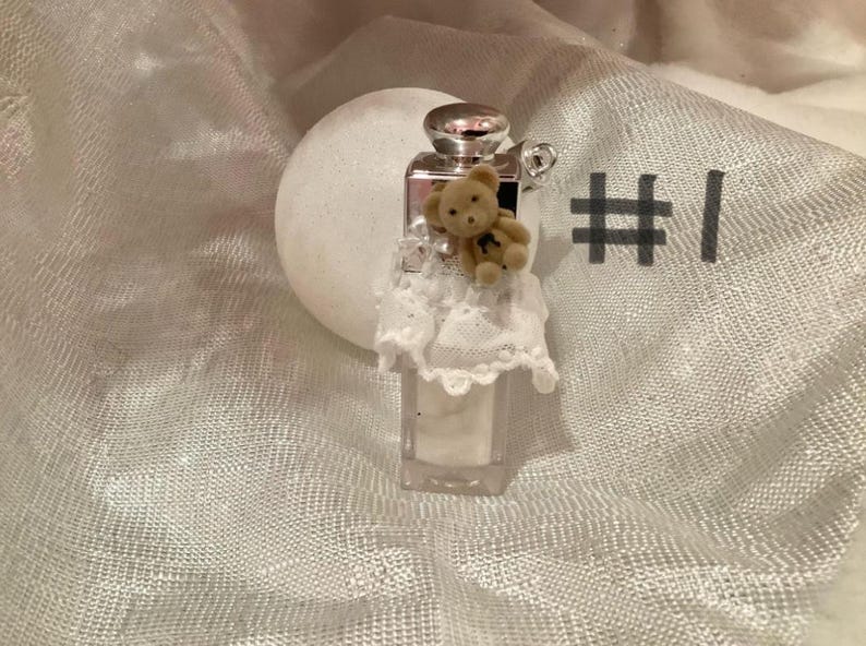 May include: A small, clear perfume bottle with a silver cap and a tiny teddy bear attached. The bottle is adorned with a white lace collar. The background is a soft, textured white fabric.