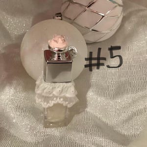 May include: A miniature perfume bottle with a silver top and a pink floral accent, adorned with a white lace skirt. The bottle is set against a backdrop of white glittery ornaments, with the number 5 written in black.