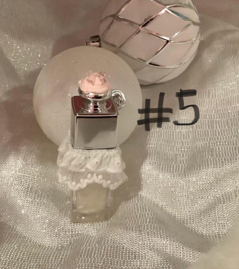 May include: A miniature perfume bottle with a silver top and a pink floral accent, adorned with a white lace skirt. The bottle is set against a backdrop of white glittery ornaments, with the number 5 written in black.